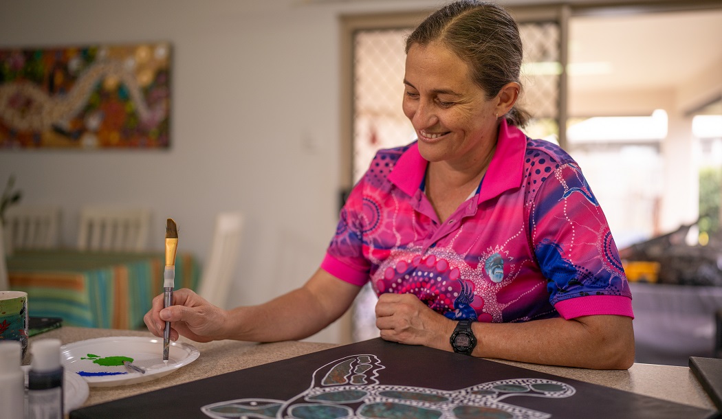 Aunty Karmen sits at the kitchen counter of her home. Her brown hair is pulled back and she is smiling as she continues working on a dot painting of a white and green sea turtle on a black background.