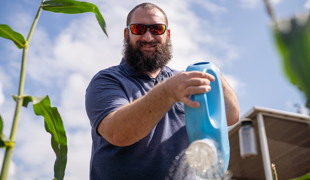 Image shows Clayton wearing sunglasses smiling down at the camera, while holding a blue plastic watering can and watering the corn in the vegetable garden.